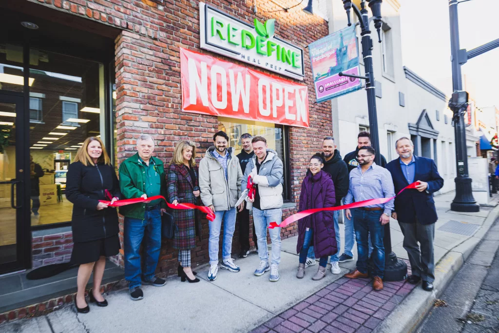 (Photo: Ron Ulip of Freshly Cut Film) Redefine Co-Owners Mark Ciabulli (fourth from left) and Matt Riss (fourth from right) prepare to cut the ribbon at their store’s grand opening. Also pictured (front row, l-r): Patchogue Chamber of Commerce President Dawn Trumbull, Patchogue Village Historian Steve Lucas, Patchogue Chamber Member Janis Thompson and Youth Outreach Coordinator Tiffany Rivera, Contour Division Manager Matthew Nieves and Patchogue Chamber Executive Director David Kennedy.