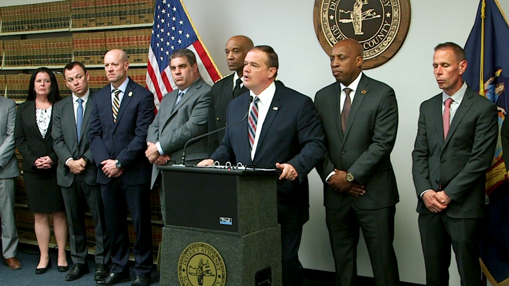 (Screen shot by Freddie Cain) Suffolk County District Attorney Ray Tierney (behind podium) speaks to the media during a press conference announcing 197 counts against 21 defendants.