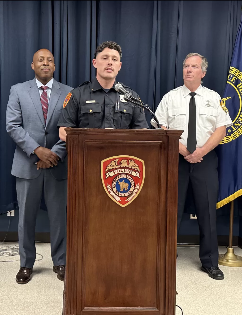 (Photo Courtesy of the SCPD) Suffolk County Police Officer Edward Pitre (standing behind podium) discusses the rescue he performed with Patchogue Fire Department firefighter Peter Feehan (right) during a June 15 press conference at SCPD headquarters in Yaphank. Also pictured is Suffolk County Police Commissioner Rodney Harrison (left).