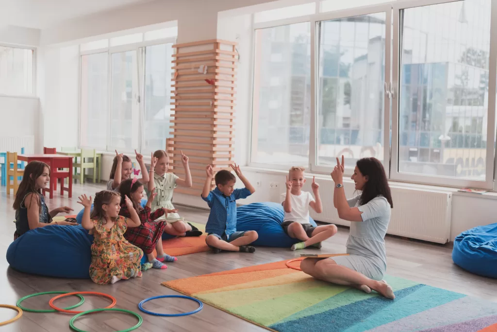 A happy female teacher sitting and playing hand games with a group of little schoolchildren