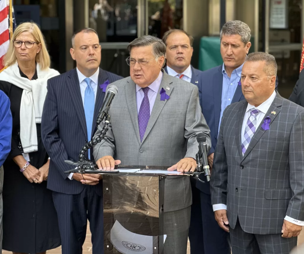 (Photo by Freddie Cain) Brookhaven Town Supervisor Ed Romaine (standing behind podium) addresses attendees in front of Brookhaven Town Hall during the “International Overdose Awareness Day” Rally on August 31. Standing next to Romaine is Brookhaven Town Councilman Michael Loguercio. Standing behind him (left to right): Town Councilman Jane Bonner, Deputy Supervisor Dan Panico, Suffolk County District Attorney Ray Tierney and Town Councilman Jonathan Kornreich.