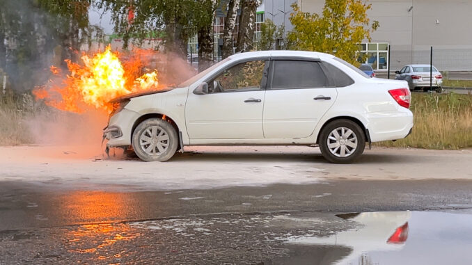 Accident. A white car in an industrial area is on fire. The engine compartment is on fire